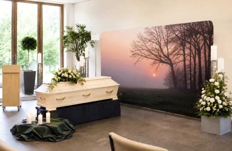 Stilvolle Trauerhallen-Dekoration mit einer Zipper-Wall im Motiv Bäume vor Sonnenuntergang neben einem hellen Holzsarg und Kerzenständern