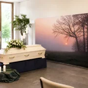 Stilvolle Trauerhallen-Dekoration mit einer Zipper-Wall im Motiv Bäume vor Sonnenuntergang neben einem hellen Holzsarg und Kerzenständern