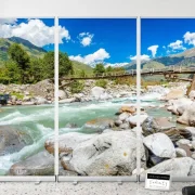 Ein kristallklarer grün-bläulicher Gebirgsbach fließt zwischen großen Felsblöcken hindurch. Eine rustikale Holzbrücke führt über das Wasser, dahinter erheben sich majestätische Berge unter blauem Himmel mit weißen Kumuluswolken. Das Motiv vermittelt Ruhe, Freiheit und hoffnungsvolle Weite.