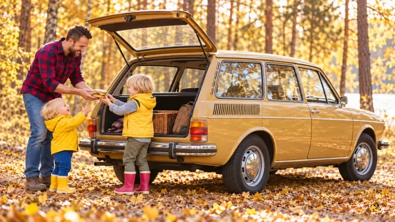 Bildbetrachtung Jahreslosung 2027 -Nach dem Waldspaziergang: gemeinsam weiter - Ein Vater hilft seinem Sohn mit roten Gummistiefeln beim Einsteigen in einen historischen VW 412 Variant; der jüngere Bruder steht neben ihnen.