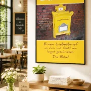 Christliches A2 Poster Liebesbrief mit gelbem historischem Briefkasten und Bibelvers aus Jeremia 31,3 in einem modernen Gemeindecafé an einer hellen Wand.