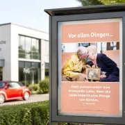 Christliches DIN A2 Poster mit 1. Petrus 4,8 in einem modernen Schaukasten vor einer Freien Evangelischen Gemeinde. Im Hintergrund sind ein modernes Gemeindegebäude und ein rotes New Beetle Cabrio zu sehen.