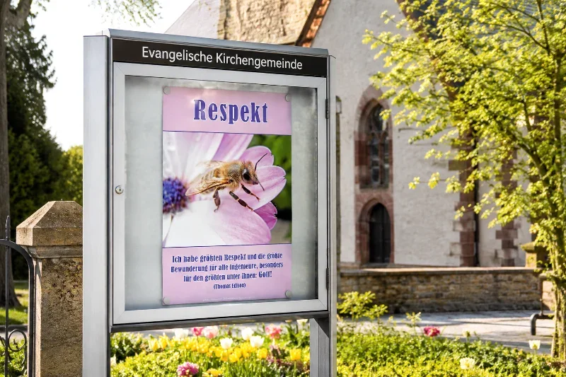 Christliches DIN-A2-Hochformat-Poster „Respekt“ mit Honigbiene und Thomas-Edison-Zitat in einem modernen Gemeindeschaukasten vor einer evangelischen Kirche im Frühling.