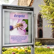 Christliches DIN-A2-Hochformat-Poster „Respekt“ mit Honigbiene und Thomas-Edison-Zitat in einem modernen Gemeindeschaukasten vor einer evangelischen Kirche im Frühling.