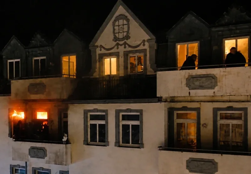 Nahaufnahme des Motivs zur Jahreslosung 2027: Ein nächtlicher Wohnungsbrand im Detail. Helle Flammen lodern auf einem Balkon, während zwei Silhouetten von Personen das Geschehen vom benachbarten Balkon aus beobachten.