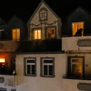Nahaufnahme des Motivs zur Jahreslosung 2027: Ein nächtlicher Wohnungsbrand im Detail. Helle Flammen lodern auf einem Balkon, während zwei Silhouetten von Personen das Geschehen vom benachbarten Balkon aus beobachten.
