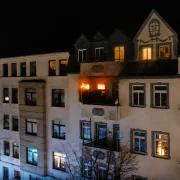 Ein mehrstöckiges Wohnhaus bei Nacht, bei dem aus einer Wohnung im Obergeschoss helle Flammen schlagen. Auf dem Nachbarbalkon beobachten zwei als Silhouetten erkennbare Personen das Feuer. Das Bild ist das Motiv der Edition Katzenstein zur Jahreslosung 2027.