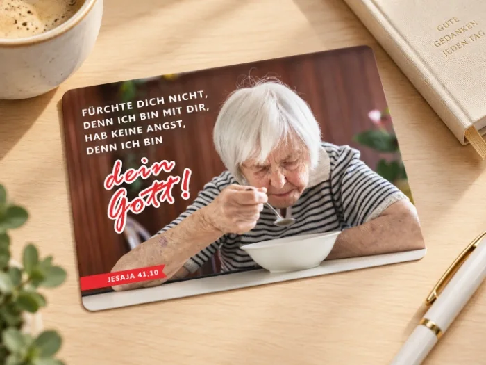 Produktfoto Postkarte Jahreslosung 2027 – Stille Mahlzeit Produktfoto der Postkarte Jahreslosung 2027 mit älterer Frau allein am Tisch auf einem stilvollen Schreibtisch