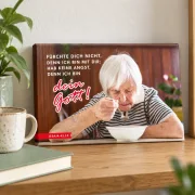 Kleines Leinwandbild zur Jahreslosung 2027 mit Seniorin beim Essen, auf einem Holzregal neben einer Tasse und einem Buch.