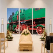 Dreiteiliges Roll-Up Display mit historischer grüner Dampflok als Hintergrund hinter einem hellen Eschenholzsarg in einer modernen deutschen Trauerhalle.