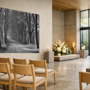 Schwarzweißes Wandbanner mit Baumallee über einem Sarg in heller Trauerhalle, umgeben von Stuhlreihen und Blumenschmuck